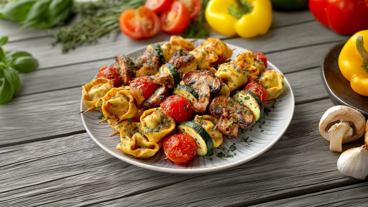 A plate of grilled tortelloni skewers. The plate is on a wooden surface. Yellow bell peppers, tomatoes, and fresh herbs are scattered around.