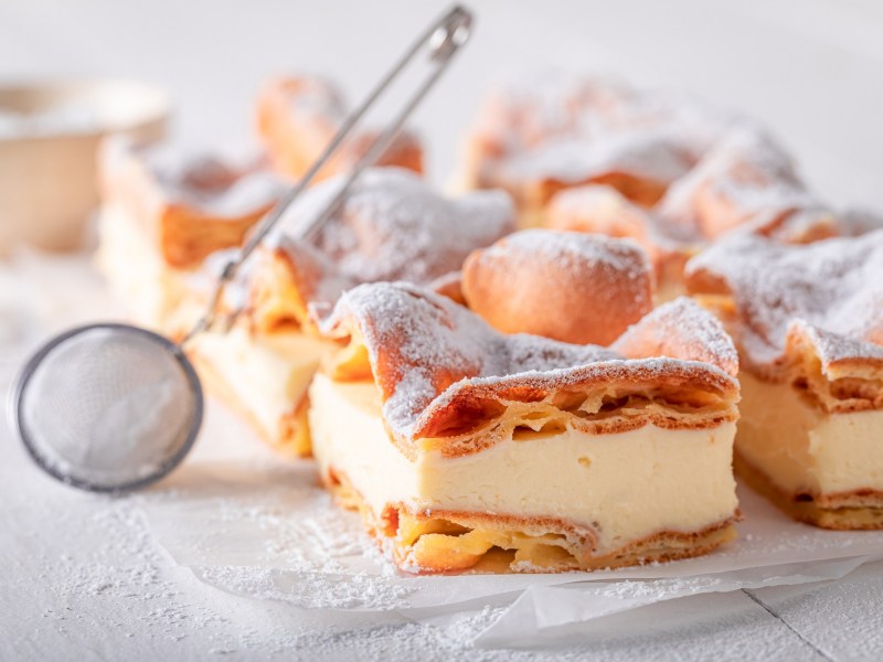 Puff pastry cake with vanilla custard on a white marble slab. On top is a sieve with which you can sprinkle the cake with powdered sugar.