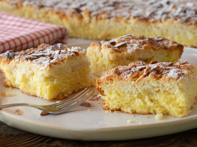 3 pieces of sheet cake butterkuchen on a plate, with the rest of the cake on a baking sheet and a kitchen towel in the background.