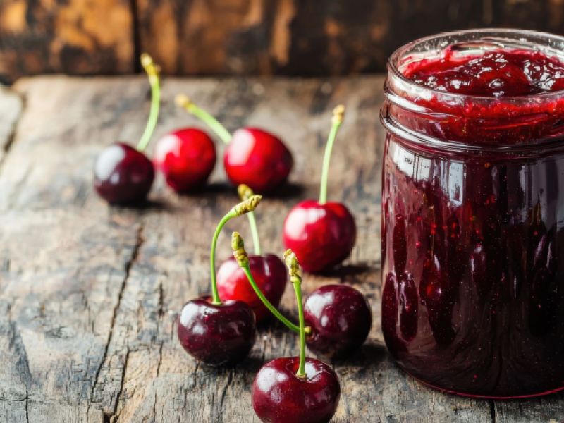 A jar of sour cherry jam, with sour cherries in the background and next to it.