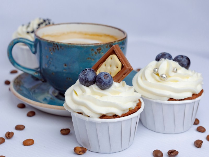 Two cappuccino cupcakes decorated with chocolate, blueberries, and sprinkles, with a blue cup of cappuccino in the background.