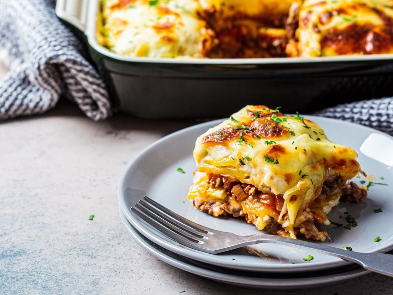 A piece of Greek Gyro Potato Casserole lies in front of a casserole dish with more casserole in it.