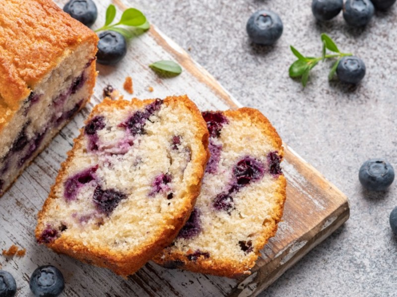 A lemon blueberry bread on a board, two slices cut off and lying next to it. Next to it are fresh blueberries and mint leaves.