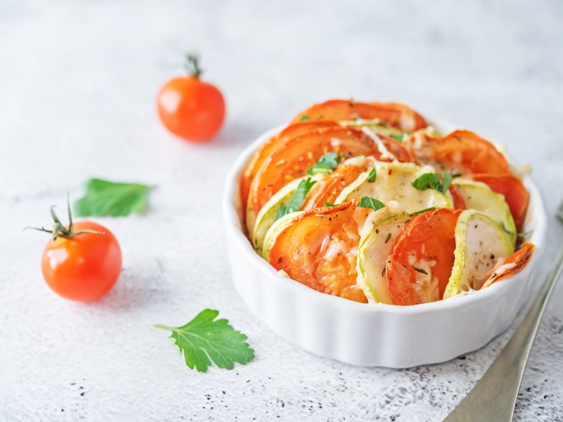 Mini vegetable gratin with tomato and zucchini in a white round baking dish, mini tomatoes next to it.